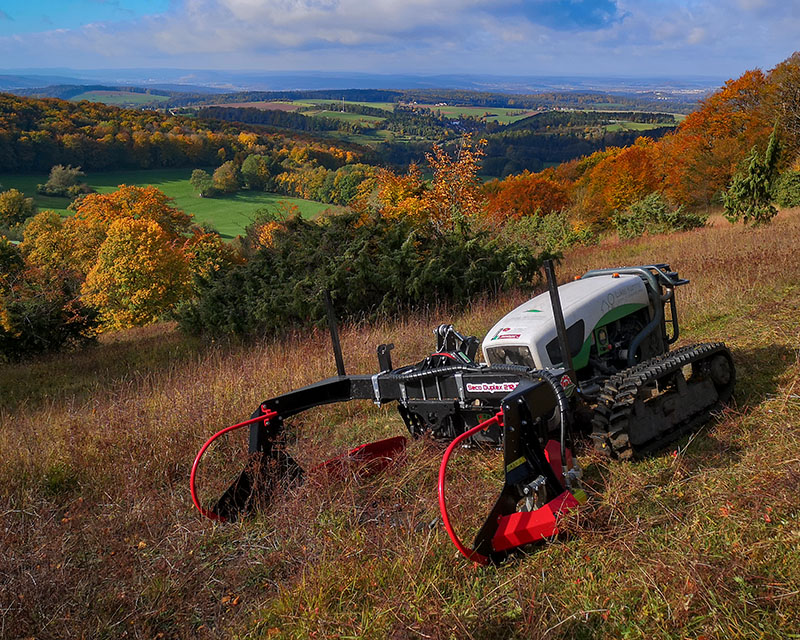 Im Vordergrund ein Geräteträger mit Kettenfahrwerk und vorgespanntem Balkenmähwerk. Er steht an einem Hang in herbstlichen Farben. Im Hintergrund das Panorama der Rhön.