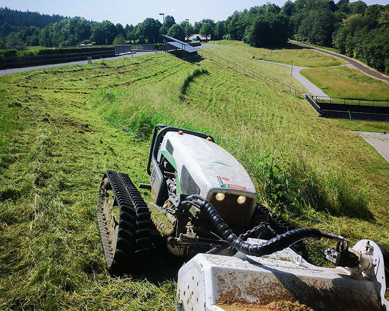 Ein ferngesteuerter Geräteträger mit Mulcher im Vordergrund mäht das Gras an einer Staumauer.