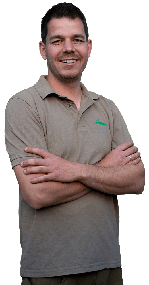Zu sehen ist Lukas Kümmel: Ein junger Mann mit schwarzen Haaren und einem Bartschatten, freundlich grinsend mit vor dem Oberkörper verschränkten Armen. Er trägt ein grau-braunes Poloshirt mit seinem Logo auf der Brust. Das Bild ist ab der Hüfte abgeschnitten.
