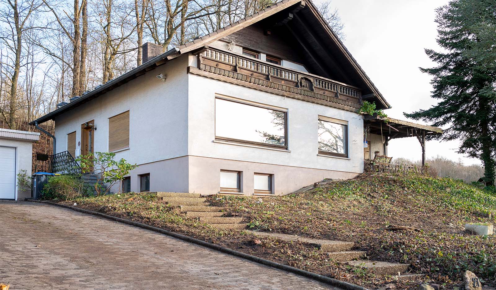Ein weiß gestrichenes Haus mit einem gepflegten Garten mit Veranda. Im Vordergrund ist die Hofeinfahrt und ein kleiner Treppenweg zur Haustür erkennbar.